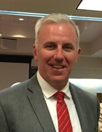 Jason Kelley, Chairman of the Board, smiles in front of a group of people.